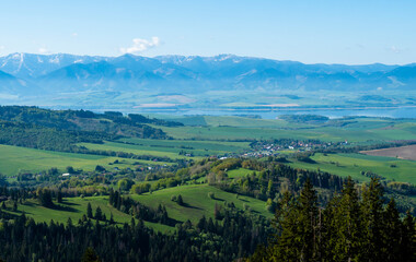 View of a village in a valley among hills with forests, agricultural fields and meadows. In the background a mountain range and a lake are visible.