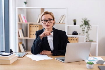 Fototapeta premium Confident adult woman in formal attire working in modern office environment. Professional focus with laptop, papers, books emphasizing business, professionalism, and productivity. Bright workspace.
