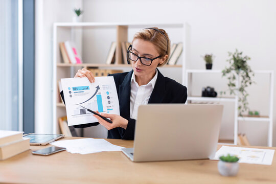 Confident businesswoman in formal outfit presenting financial documents at office desk with laptop. Office plant decor is visible in background