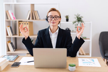 Adult woman meditating at office desk in lotus pose wearing glasses and suit promoting work-life balance and mindfulness during break period enhancing productivity and maintaining demeanor.