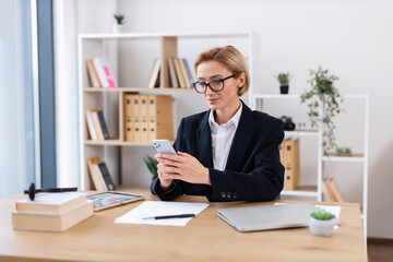 Businesswoman wearing glasses working at office desk using smartphone surrounded by work tools....