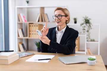 Smartly dressed businesswoman seated at desk using smartphone with happy expression. Office features organized bookshelf, papers, and plants creating professional environment.