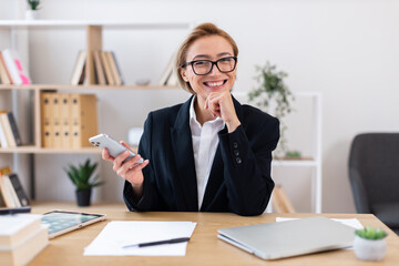 Confident adult woman in formal clothing holding smartphone sitting at desk in office. Workplace features documents, laptop, modern decor. Symbolizes professionalism, communication success in business