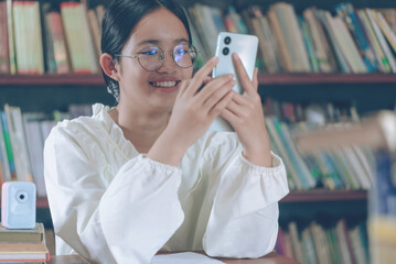 Young girl with glasses watching social media online on the phone bookshelf background.