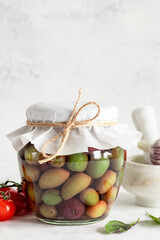 A glass jar with various pickled olives in brine stands on a white table.