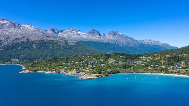 Aerial view of a turquoise lake, lush green shore with houses, & snow-capped mountain range under blue sky