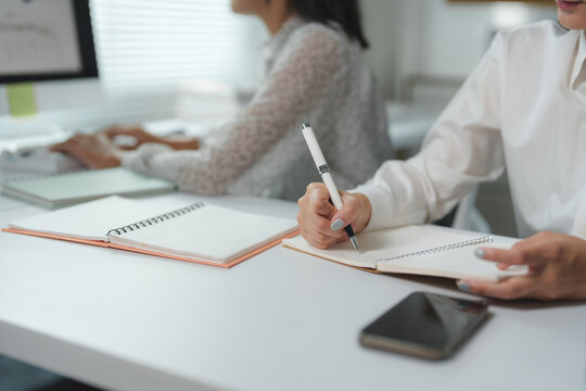 Businesswoman taking notes in spiral notebook during corporate meeting, with colleague working on computer in background, showcasing collaborative office environment