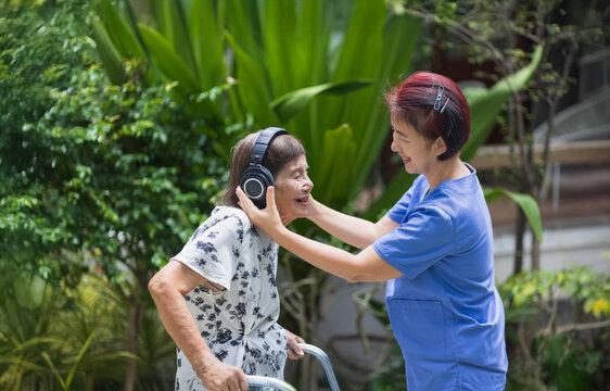 Asian senior woman listening music by headphone with caregiver in backyard.
