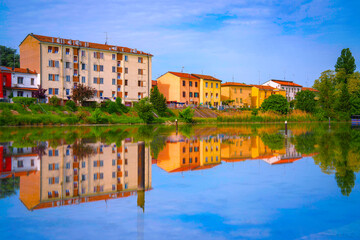 Fototapeta premium Tranquil Po River summer landscape in Ferrara, Emilia-Romagna, Italy, with colorful traditional houses reflected on the calm water—where nature and heritage mirror each other in peace