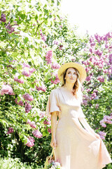 Fototapeta premium Young Woman In Summer Hat Enjoying The Fragrance Of Lilac Blooms On Sunny Day. lady in a straw hat and dress smelling lilac flowers in spring sunlit garden.