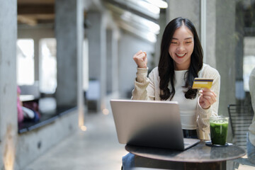 Young asian woman holding a credit card and celebrating a successful online transaction, expressing...