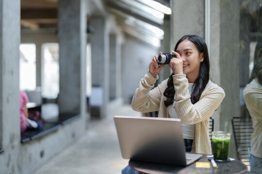 Asian female photographer taking pictures with a digital camera in a modern cafe, she's working with her laptop and enjoying a healthy green juice