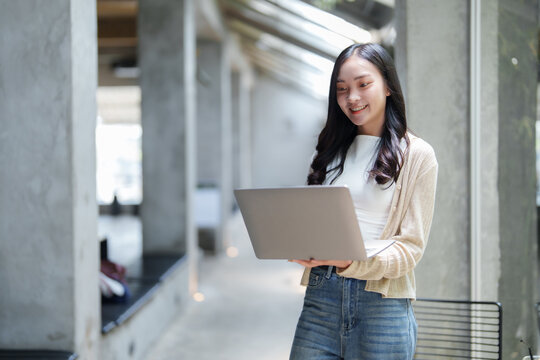 Smiling asian freelancer standing and using laptop in modern office space, enjoying remote work and flexible schedule, benefiting from digital nomad lifestyle