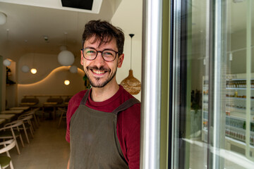 Portrait of a young entrepreneur smiling and welcoming customers at the door of his coffee shop or...