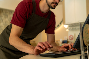 Shopkeeper wearing an apron is typing on the keyboard of a POS terminal, processing customer orders...