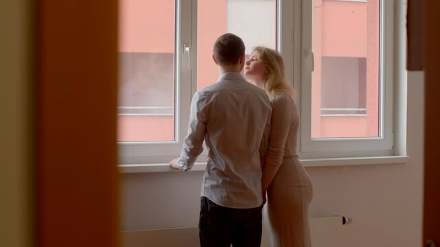 A couple stands by a large window, gazing outside together, sharing a moment of tranquility