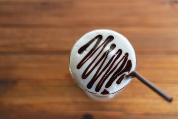op view of a creamy iced beverage with thick foam and artistic chocolate syrup drizzle, served with a brown straw on a rustic wooden table.
