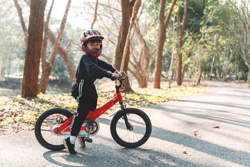 Naklejka premium Young child with helmet learning to ride a red bicycle on a peaceful forest path in warm morning sunlight. Focused and determined expression.