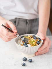 Bowl of granola with greek yogurt, kiwi and blueberries and spoon in hands of woman on white table.