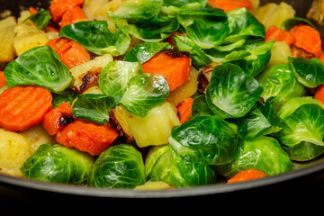 Healthy comfort food preparation captured mid-cook, with brussel sprouts caramelizing, potatoes browning and carrots glistening in minimal oil in commercial-grade skillet. 