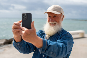 A happy elderly man with a white beard, wearing a beige cap and denim shirt, takes a selfie with his smartphone near the ocean, smiling as he captures the moment