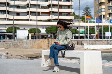 A young woman with curly dark hair, wearing sunglasses, a green jacket, and jeans, sits on a white bench near the beach, looking at her smartphone with a black shoulder bag beside her