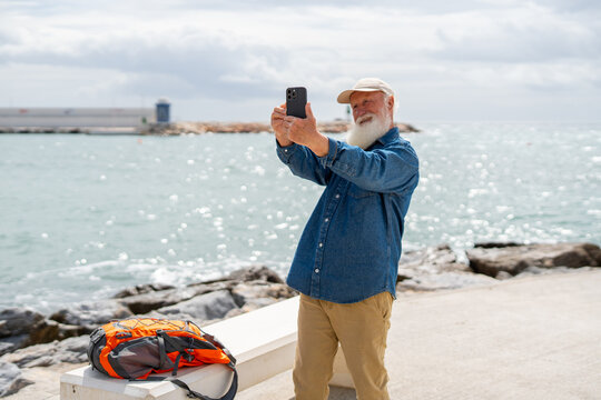 A cheerful elderly man with a white beard, wearing a beige cap and denim shirt, takes a selfie with his smartphone near the ocean, with his orange backpack resting on a white bench - Powered by Adobe