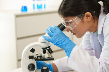 Asian female student wearing lab coat and safety goggles closely examines specimen under microscope...