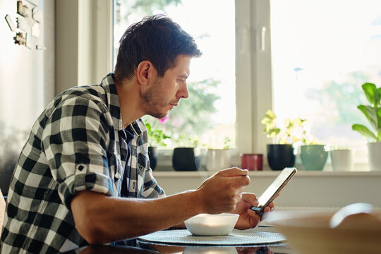 Man in checkered shirt eats from bowl at kitchen table and watching video on his smartphone. Man distracted by phone while eating breakfast at home. Social media and online content addiction