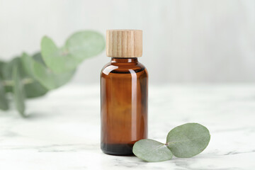 Bottle of essential oil and eucalyptus leaves on white marble table, closeup