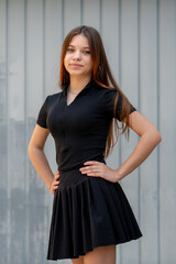 A young girl in black clothing stands against a grey wall, posing confidently with her hands on her...