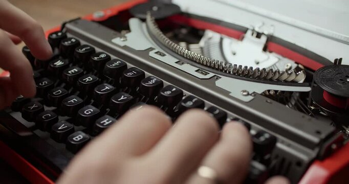 Close-up shot of hands typing on a retro red typewriter. Black and silver keys, fingers curled mid-action, wooden surface, paper in background. Elevated angle with depth.