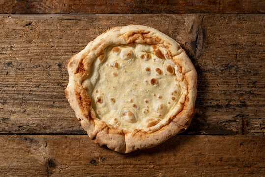 Pre-baked round pizza base placed on rustic wooden surface. White dough, ready to be topped and baked, in a homemade or artisanal context.