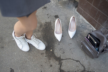 women's feet in white sneakers standing on the asphalt. There are white high-heeled shoes and a black bag nearby.The business lady changed her shoes
