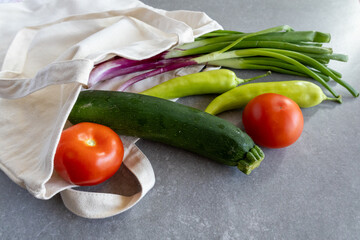 A reusable cotton shopping bag lies on a gray countertop with fresh vegetables spilling out.