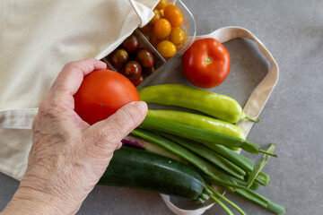 A hand holds a tomato above fresh vegetables spilling from a reusable canvas bag.