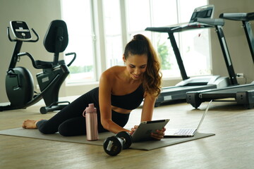 Beautiful Latin woman with a good figure sitting in a gym looking at a laptop to practice yoga exercise for health and weight loss in a fitness center.