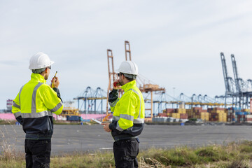 Two port authority officers in high-visibility jackets and white helmets communicate via walkie-talkies while overseeing operations at a shipping port with large cranes and stacked containers.