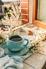 Blue cup with white clouds filled with tea or coffee, surrounded by cherry blossoms on a soft pastel fabric. Dreamy spring morning scene by the window