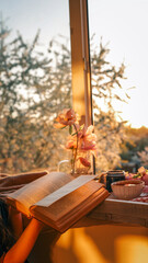 Person reading a book by a cozy window at sunset, with magnolia flowers, a black candle, and a cup of tea on a wooden tray. Warm golden light creates a peaceful, serene mood