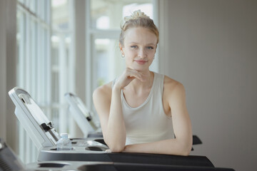 Beautiful woman is exercising on a treadmill with water placed between her. She stands and takes a portrait and smiles happily.