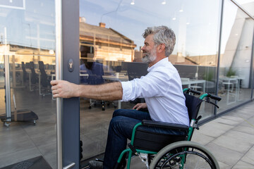 Gray-haired bearded mature man on a wheelchair opening the door to the office