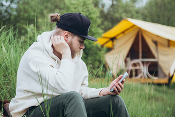 Young man uses cell phone, sitting near the tent during camping.