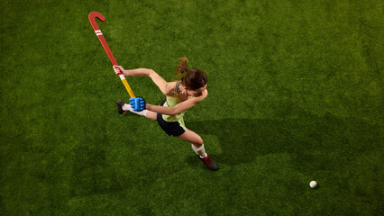 Aerial view photo of sportswoman swings hockey stick with dynamic energy on green grass field against white studio background, capturing motion in game moment. Concept of sport, championship, movement