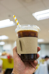 Iced americano coffee in plastic galls with brown paper sleeve cup and paper straws striped golden in office on hand