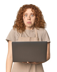 Young Caucasian redhead woman with laptop shrugs shoulders and open eyes confused.
