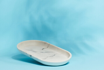 An empty, oval shaped soap dish made of white marble with gray veining sits on a light blue background