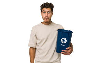 Young caucasian man holding a paper trash for recycle shrugs shoulders and open eyes confused.