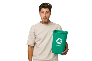 Young caucasian man holding a glass trash isolated shrugs shoulders and open eyes confused.