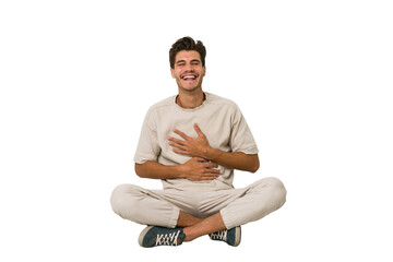 Young caucasian man sitting on the floor isolated on white background laughs happily and has fun keeping hands on stomach.